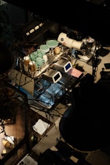 A top-down view of a coffee preparation station that includes a high-end espresso machine, milk frothing equipment, and various coffee cups and saucers. The area's lighting appears dim, suggesting a cozy, intimate setting. There are various objects such as cups, paper coffee cups, and a small white towel.