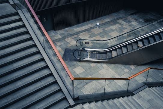 A modern architectural setting features a wide staircase with a metal handrail on the left and a sleek escalator to the right. The ground is made of gray stone tiles, and the surroundings are composed of clean lines and minimalist design elements. Both the staircase and the escalator are unoccupied.
