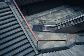A modern architectural setting features a wide staircase with a metal handrail on the left and a sleek escalator to the right. The ground is made of gray stone tiles, and the surroundings are composed of clean lines and minimalist design elements. Both the staircase and the escalator are unoccupied.