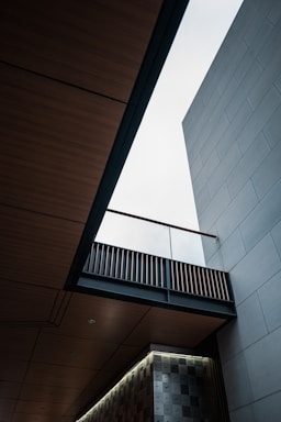 An architectural scene featuring a modern building with clean lines and geometric shapes. The view looks upward, showcasing a bright sky through an opening between wooden and concrete elements. The design includes a patterned wall and an overhanging bridge-like structure with slats.