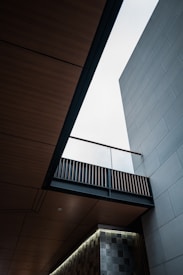 An architectural scene featuring a modern building with clean lines and geometric shapes. The view looks upward, showcasing a bright sky through an opening between wooden and concrete elements. The design includes a patterned wall and an overhanging bridge-like structure with slats.