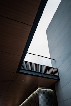 An architectural scene featuring a modern building with clean lines and geometric shapes. The view looks upward, showcasing a bright sky through an opening between wooden and concrete elements. The design includes a patterned wall and an overhanging bridge-like structure with slats.