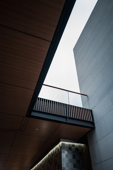 An architectural scene featuring a modern building with clean lines and geometric shapes. The view looks upward, showcasing a bright sky through an opening between wooden and concrete elements. The design includes a patterned wall and an overhanging bridge-like structure with slats.