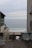 A serene beachfront scene framed between two buildings. The ocean is calm with gentle waves hitting the sandy shore. There are two white chairs in the foreground on a terrace, positioned to enjoy the view. The sky is overcast with a hint of soft sunlight breaking through, creating a peaceful atmosphere.