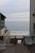 A serene beachfront scene framed between two buildings. The ocean is calm with gentle waves hitting the sandy shore. There are two white chairs in the foreground on a terrace, positioned to enjoy the view. The sky is overcast with a hint of soft sunlight breaking through, creating a peaceful atmosphere.