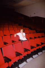 A person sits alone in a dimly lit theater with rows of empty red seats. The individual is wearing a white shirt and appears to be in a relaxed posture with one arm resting on the back of a seat.