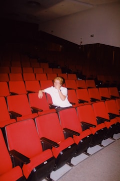 A person sits alone in a dimly lit theater with rows of empty red seats. The individual is wearing a white shirt and appears to be in a relaxed posture with one arm resting on the back of a seat.