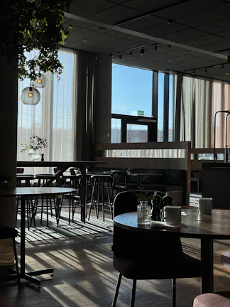 Sunlight streaming through large windows onto rustic wooden tables with steaming coffee cups.