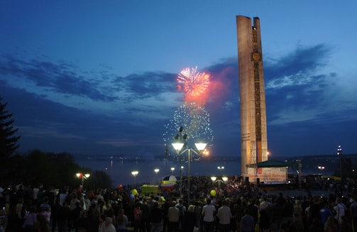A large crowd gathers at an outdoor event during twilight, with vivid fireworks bursting in the sky. A tall monument towers over the scene, lit by artificial lights. The darkening sky creates a dramatic backdrop, accentuating the warm glow from the lights and fireworks.