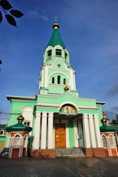 A large, ornate church features a tall green and white bell tower topped with a gold cross. The building has decorative arches, columns, and icons above the wooden entrance door. The architecture is highlighted by golden domes with Orthodox crosses.
