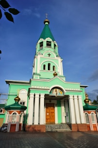 A large, ornate church features a tall green and white bell tower topped with a gold cross. The building has decorative arches, columns, and icons above the wooden entrance door. The architecture is highlighted by golden domes with Orthodox crosses.