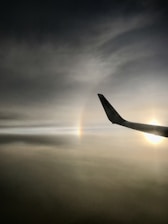 A dark, moody sky with a faint rainbow visible and the silhouette of an aircraft wing. The wing displays the logo of Ryanair and a bright sun can be seen on the right side.