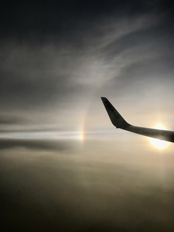 A dark, moody sky with a faint rainbow visible and the silhouette of an aircraft wing. The wing displays the logo of Ryanair and a bright sun can be seen on the right side.