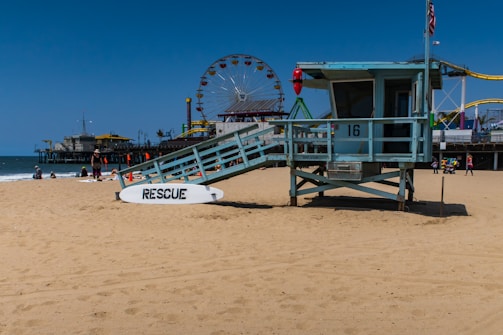 A sandy beach with a lifeguard station numbered 16. A white rescue board leans against the station. In the background, a pier extends into the ocean with various amusement park rides, including a large Ferris wheel. Several people are relaxing and walking on the beach.