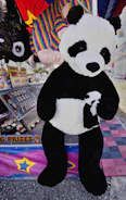 A large panda plush toy is displayed at a carnival booth. The booth features a sign advertising big prizes and colorful plush toys in the background, including a rainbow-colored toy. The environment is festive, with vibrant colors and promotional materials for winning prizes.