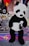 A large panda plush toy is displayed at a carnival booth. The booth features a sign advertising big prizes and colorful plush toys in the background, including a rainbow-colored toy. The environment is festive, with vibrant colors and promotional materials for winning prizes.