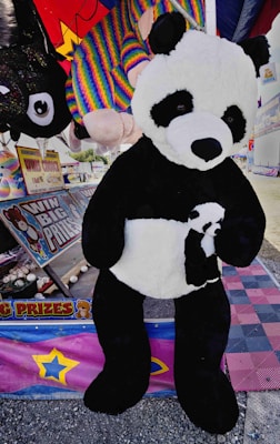 A large panda plush toy is displayed at a carnival booth. The booth features a sign advertising big prizes and colorful plush toys in the background, including a rainbow-colored toy. The environment is festive, with vibrant colors and promotional materials for winning prizes.