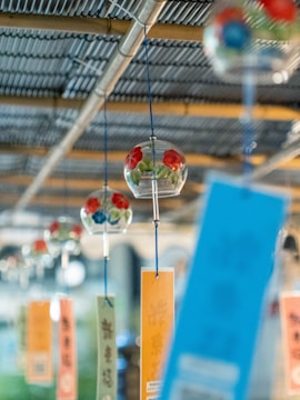 Transparent glass wind chimes with colorful floral patterns hang in a row, accompanied by rectangular papers of various colors. The background shows a blurred metallic ceiling with wooden beams.