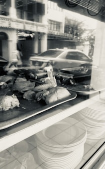 Pieces of cooked chicken are displayed on metal trays in a window, with a reflection showing a street and a parked car. Beneath the trays, stacks of white plates are visible on a shelf. The overall setting appears to be a restaurant or food shop.