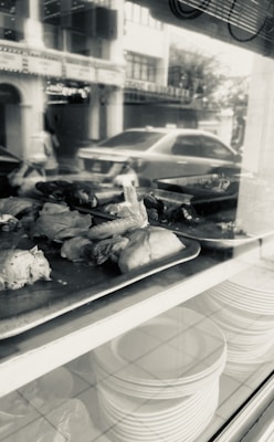 Pieces of cooked chicken are displayed on metal trays in a window, with a reflection showing a street and a parked car. Beneath the trays, stacks of white plates are visible on a shelf. The overall setting appears to be a restaurant or food shop.