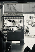 A monochrome image of a hawker stall selling Hainan chicken rice. Inside the stall, chicken pieces are hung up for display. Outside, a street view with motorbikes and cars can be seen.