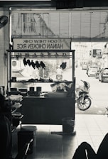 A monochrome image of a hawker stall selling Hainan chicken rice. Inside the stall, chicken pieces are hung up for display. Outside, a street view with motorbikes and cars can be seen.