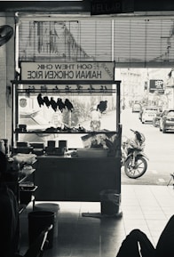 A monochrome image of a hawker stall selling Hainan chicken rice. Inside the stall, chicken pieces are hung up for display. Outside, a street view with motorbikes and cars can be seen.