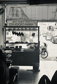 A monochrome image of a hawker stall selling Hainan chicken rice. Inside the stall, chicken pieces are hung up for display. Outside, a street view with motorbikes and cars can be seen.