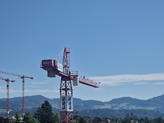 A large red construction crane with the word 'WOLFF' on its arm is seen against a backdrop of clear blue skies and distant green mountains. To the left, there are two additional cranes visible, partially obscured by the foreground crane. The surrounding area includes trees and buildings, suggesting a construction site in a mountainous region.