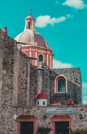 A historic stone church with a large, ornate dome is set against a bright blue sky. The architecture features intricate details with reddish-pink accents and tall, arched windows. A sign on the wall reads 'Parroquia Santa María de la Asunción.'