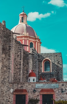 A historic stone church with a large, ornate dome is set against a bright blue sky. The architecture features intricate details with reddish-pink accents and tall, arched windows. A sign on the wall reads 'Parroquia Santa María de la Asunción.'