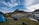 A picturesque mountain landscape featuring a blue tent set up on a grass field near a tranquil lake. Snow patches cover parts of the rocky mountain in the background. A pair of hiking boots and a backpack are placed on the grass, symbolizing an outdoor adventure.