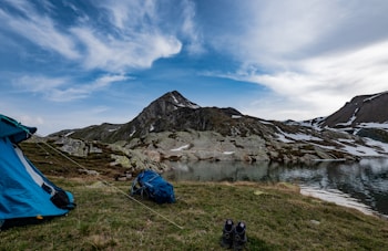 A picturesque mountain landscape featuring a blue tent set up on a grass field near a tranquil lake. Snow patches cover parts of the rocky mountain in the background. A pair of hiking boots and a backpack are placed on the grass, symbolizing an outdoor adventure.