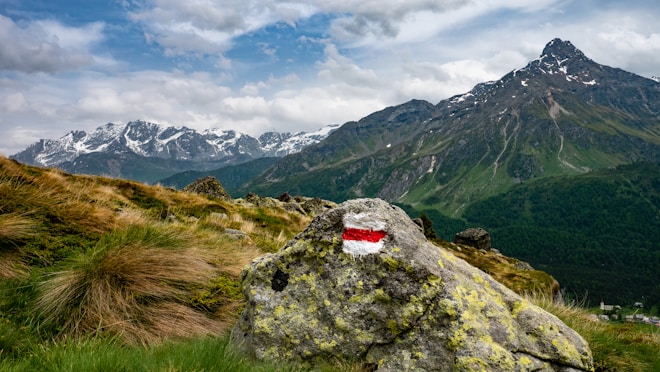 A scenic mountain landscape featuring a prominently marked rock in the foreground with a red and white stripe. The background showcases rugged mountain peaks with patches of snow under a partly cloudy sky. Lush green grass and small shrubs cover the terrain, adding a sense of vibrant nature.