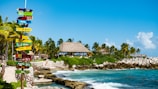 A tropical beach scene with a colorful signpost pointing towards various countries. There is a thatched-roof hut in the background surrounded by lush palm trees. Crystal-clear blue water and rocky coastline create a picturesque seaside atmosphere.