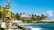 A tropical beach scene with a colorful signpost pointing towards various countries. There is a thatched-roof hut in the background surrounded by lush palm trees. Crystal-clear blue water and rocky coastline create a picturesque seaside atmosphere.