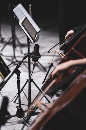 Musicians are seen playing string instruments, possibly cellos, with a music stand and microphone in view. The scene suggests a rehearsal or performance setting with a focus on the instruments and the musicians' hands.