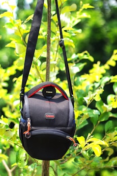 A black camera bag with red accents hangs from a branch amidst vibrant green leaves. The bag is branded with Canon logos and features a shoulder strap. Sunlight casts a bright and lively ambiance on the foliage.