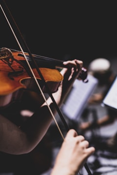 A close-up of a person playing a violin, with focus on the violin strings and bow. The background is blurred, suggesting a musical performance or practice session.