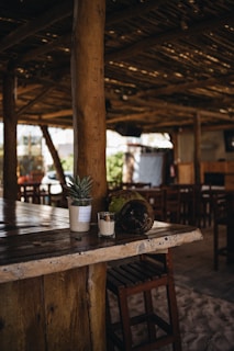A rustic, wooden bar setting with a wooden beam and countertop. There is a small potted plant and a coconut placed on the counter, along with a glass with a tealight. The environment has wooden chairs and tables, with sandy flooring, suggesting an outdoor or beachside bar.