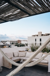 A cozy rooftop terrace featuring a comfortable L-shaped sofa with light-colored cushions sits against a white wall. A hammock is stretched diagonally across the space under a rustic wooden pergola that casts interesting shadows. The sky is clear with a gentle blue tone, and various modern buildings extend into the distance.
