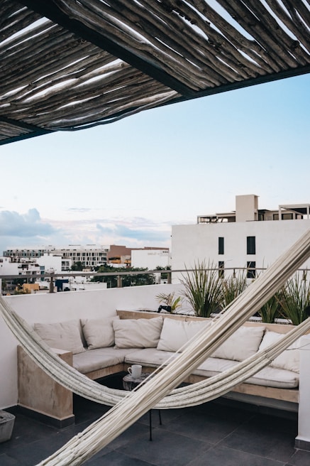 A cozy rooftop terrace featuring a comfortable L-shaped sofa with light-colored cushions sits against a white wall. A hammock is stretched diagonally across the space under a rustic wooden pergola that casts interesting shadows. The sky is clear with a gentle blue tone, and various modern buildings extend into the distance.