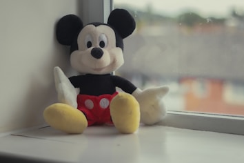A plush toy resembling a well-known cartoon character sits on the edge of a windowsill. The toy has large black ears, a white face with black eyes, red shorts with white buttons, and yellow shoes. The background outside the window is blurred, suggesting a view of rooftops under a cloudy sky.