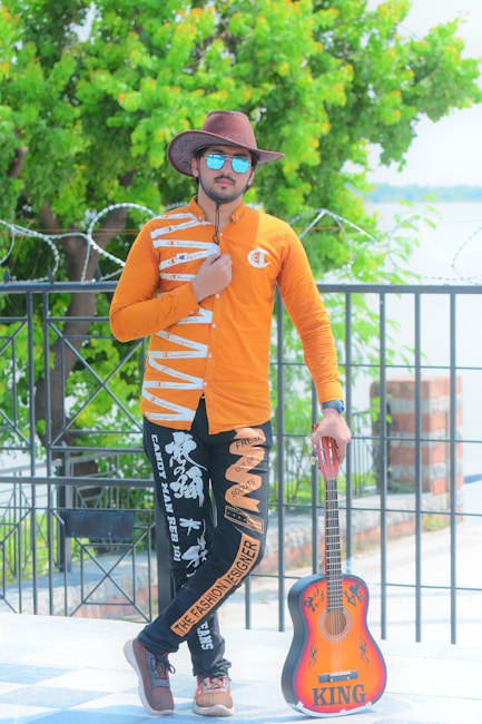 A person wearing a cowboy hat, sunglasses, and a bright orange shirt with unique designs stands confidently near a fence. The individual is posing with one hand on their chest and the other resting on a colorful guitar labeled 