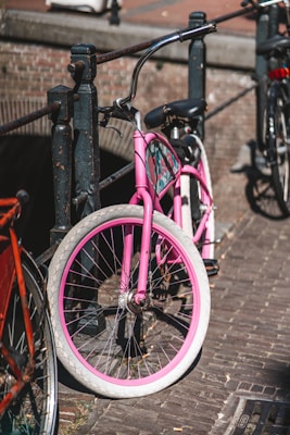 A pink bicycle is parked against a black iron railing on a cobblestone street. The bicycle has white tires with pink rims and is positioned near a bridge over a canal. Another bicycle with red elements is partially visible in the foreground, and there's a hint of a brick wall in the background.