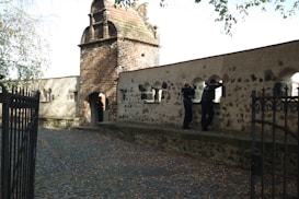A cobblestone pathway leads to a historic stone structure with a small tower and arched openings. Three people, two of whom appear to be wearing police uniforms, are standing near the structure's openings, with one person looking through one of the arches. The scene is shaded by overhanging tree branches.
