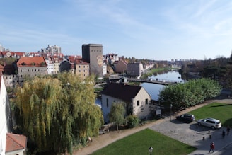 An urban riverfront view featuring charming, historic buildings with red roofs lining the riverbank. A pedestrian bridge stretches over the river, surrounded by greenery including trees and grassy areas. A few people and cars are visible, contributing to a peaceful, lively atmosphere under a clear blue sky.