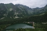 A serene mountain landscape with towering, rugged peaks partially shrouded in clouds. Below the mountains lies a dense forest surrounding a tranquil, reflective lake. A small cluster of buildings sits at the water's edge, nestled in the greenery.