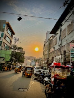 A bustling street scene at sunset with multiple auto rickshaws and a few pedestrians. The street is lined with buildings, some adorned with signage. A large, historic archway is visible in the distance. Power lines crisscross above, and there are vendors with colorful carts on the side.