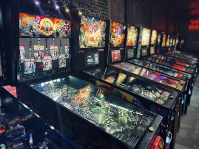 A row of colorful pinball machines is displayed against a dimly lit interior with brick walls. Each machine features bright, intricate artwork depicting various themes and characters. Neon lights reflect off the glass surfaces, enhancing the vibrant and lively atmosphere.